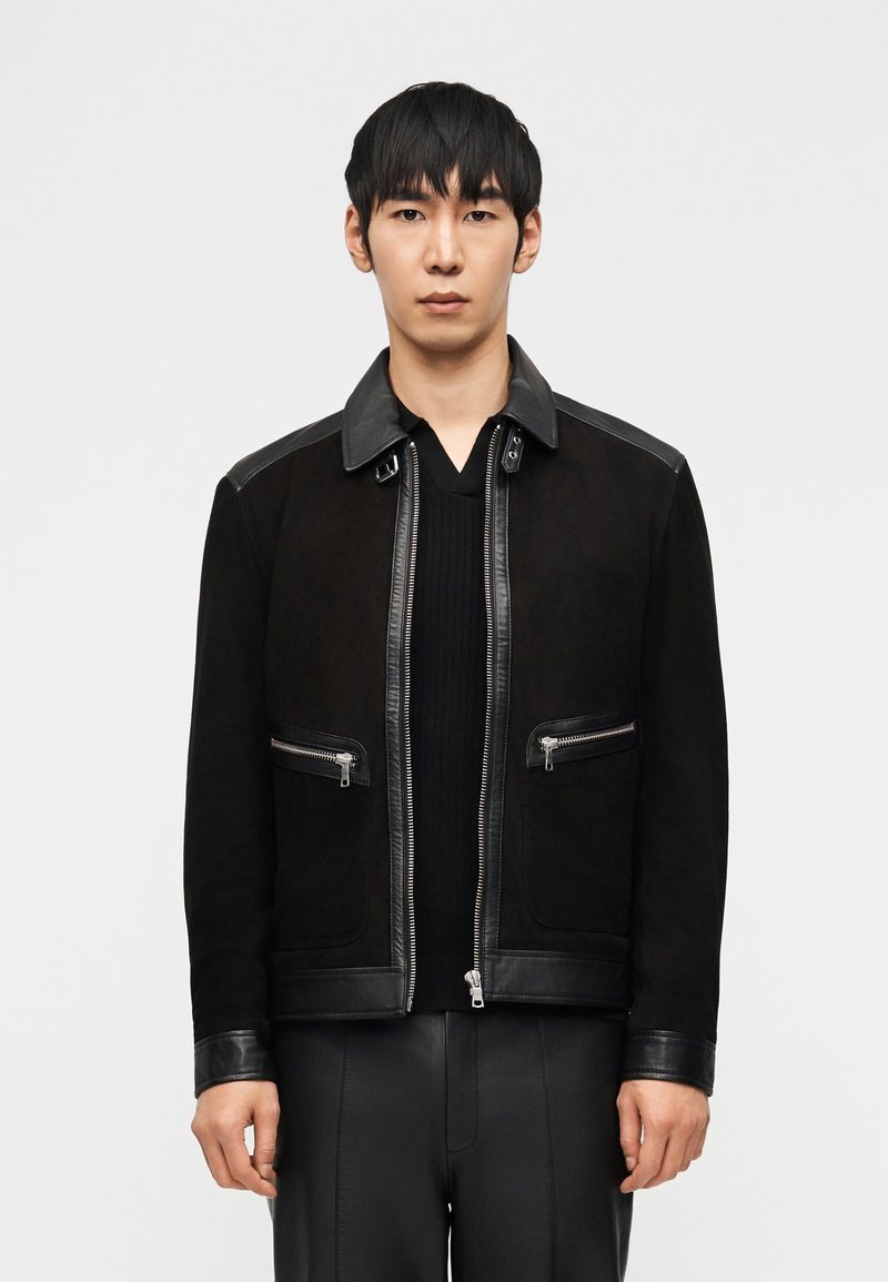 Άνδρας που φοράει μαύρο μπουφάν με δερμάτινες λεπτομέρειες και τσέπες με φερμουάρ πάνω από μαύρο ριμπ πουλόβερ με V-λαιμόκοψη, στέκεται μπροστά σε λευκό απλό φόντο.