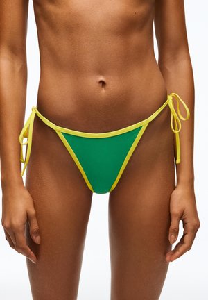 Kobieta w zielonych majtkach bikini z żółtym obszyciem i troczkami po bokach, pokazana od połowy torsu do górnej części ud na białym tle.