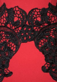 Czerwona tkanina z misterną czarną koronką, która pokrywa wzory kwiatowe i okrągłe motywy wzdłuż dekoltu, tworząc wyrazisty kontrast.