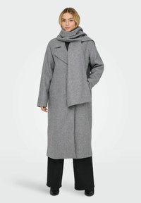 Cappotto di lana grigio con design avvolgente, colletto profondo e sciarpa abbinata. Tessuto texture. Indossato sopra pantaloni neri, che completano il colore del cappotto.