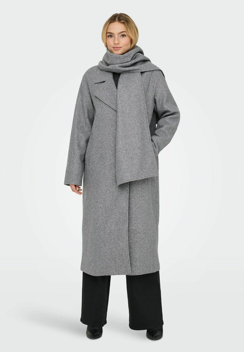 Manteau en laine grise avec un design enveloppant, un grand revers et une écharpe assortie. Tissu texturé. Porté sur un pantalon noir, complétant la couleur du manteau.