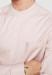 Ljusrosa skjorta med mandarin-krage, försedd med vita knappar och en bröstficka. Slät textur och figurnära design.