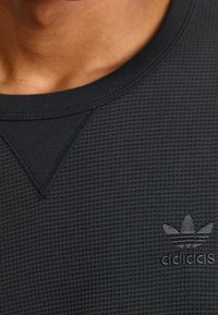 Svart långärmad tröja i våffelstruktur med rund halsringning, med en subtil mörkgrå Adidas-logotyp på vänster bröst.