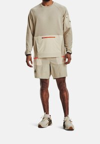 Uomo che indossa una felpa beige a maniche lunghe con tasca frontale, pantaloni corti coordinati e scarpe da ginnastica beige con accenti neri e marroni.