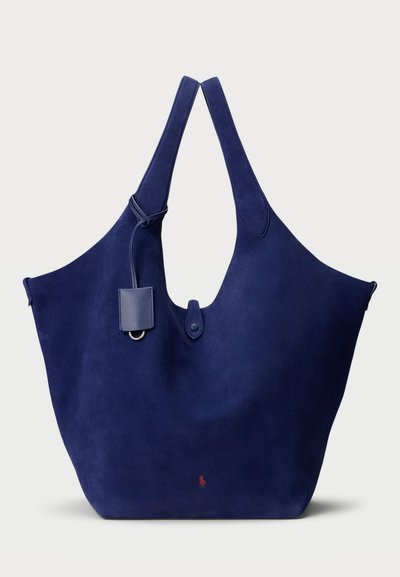 Sac fourre-tout en suédine bleu marine avec une forme courbe large, deux poignées, et une étiquette amovible. Présente un petit logo rouge à l'avant.