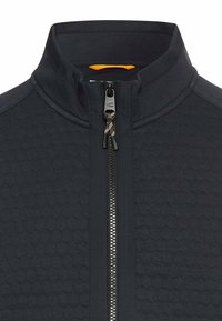 Close-up van een donkerblauwe ritsjack met een hexagonaal textuurpatroon en een hoge kraag, met een zichtbare gele voering.