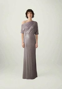 Lühikeste kurruliste juustega naine, kes kannab pikka, õlavarrelt avatult, sädelevat halli-lilla kleiti, seismas siledal heledal taustal.