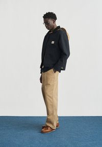 Uomo con giacca nera, pantaloni color khaki, scarpe marroni e occhiali, in piedi di lato su un tappeto blu con un cappello beige appeso sulla schiena.