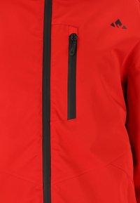 Rode softshell jas met zwarte ritssluiting en accenten op de zakken. Gladde textuur met hoekige naaddetails en een minimalistisch logo op de borst.