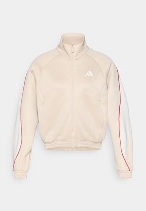 Beige jakke med glidelås og høy krage, hvite og røde sideakcenter, ribbestrikkede mansjetter og kant. Har en liten logo på brystet og to lommer.