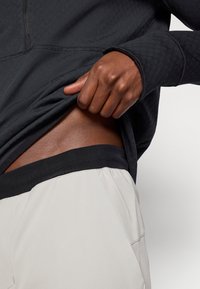 Fekete cipzáras kapucnis pulóver texturált anyagból, részben felhúzva, hogy felfedje a kontrasztos fekete derékpántot a világos bézs nadrágon. Zökkenőmentes bőr látható.