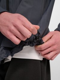 Ręce regulują ściągacz na dole ciemnej kurtki z długim rękawem, na białej koszuli i czarnych spodniach.