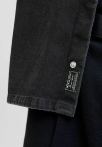 Black denim jacket collar with a silver snap button and a black label featuring white text. Smooth texture and clean stitching details.