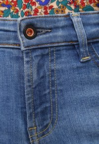 Denimjeans har ett blått tyg med en mjuk textur, en kopparfärgad knapp och kontrasterande gula sömmar längs sömmarna.