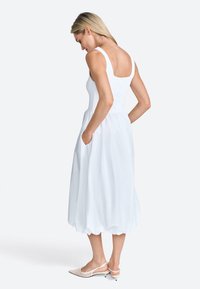 Witte mouwloze jurk met een aansluitend lijfje en een vloeiende, enkel-lange rok; heeft zijzakken en een gathered zoom. Eindigt boven de enkel met puntige schoenen.