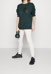 Felpa verde scuro con logo Adidas nero, abbinata a pantaloni jogger color crema con strisce laterali bianche e scarpe sportive nere.
