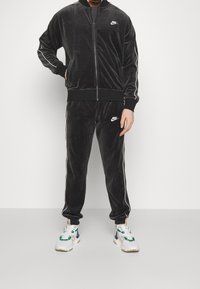 Černá semišová tepláková souprava s bílými prvky. Bunda se zipem a odpovídající kalhoty mají postranní pruhy a logo Nike. Outfit doplňují tenisky.