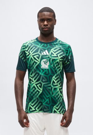 Mężczyzna ubrany w zieloną koszulkę piłkarską Meksyku z geometrycznym wzorem i logo Adidas, stojący na tle jasnego, jednolitego tła.
