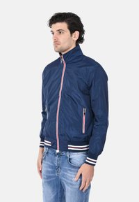 Giacca in nylon blu navy con colletto alto, chiusura con zip frontale e due tasche laterali. I polsini e l'orlo presentano accenti a strisce bianche e rosse.