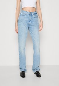 Jeans de tiro alto de color azul claro con un diseño de pierna recta y bolsillos frontales visibles. Se combinan con zapatos negros sin cordones con detalle de cadena.