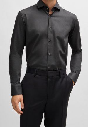 Hombre con una camisa gris oscuro ajustada abotonada, metida dentro de un pantalón de vestir negro, con una mano en el bolsillo.