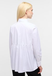 Camicia bianca a maniche lunghe con colletto alto, design con schiena arricciata, texture liscia e polsini con bottone. Stile minimalista e vestibilità sartoriale.