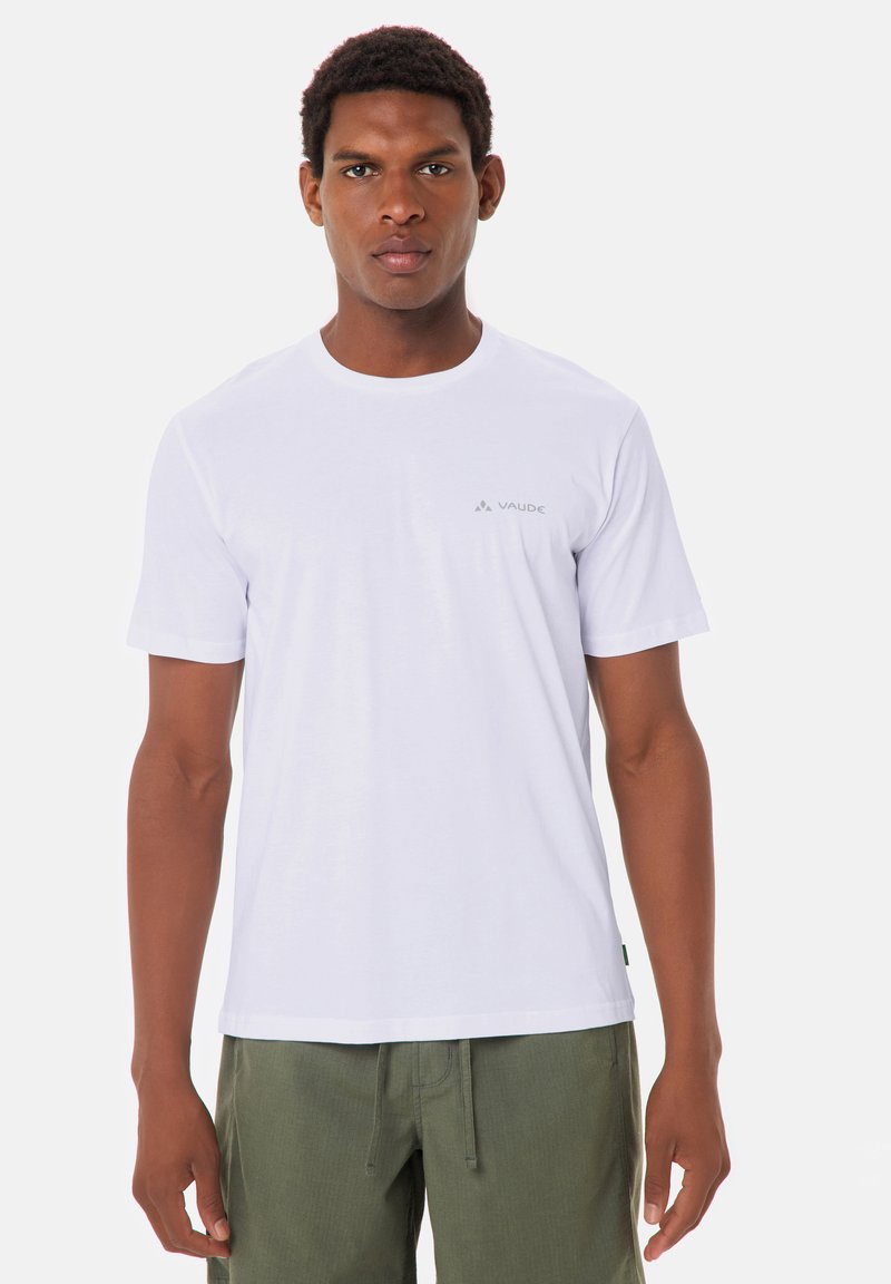 Giovane uomo che indossa una maglietta bianca semplice di Vaude e pantaloni verde oliva, in piedi contro uno sfondo chiaro e uniforme.