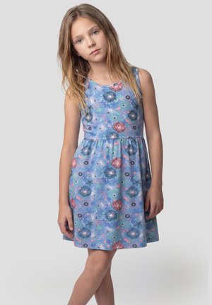 Niña joven con cabello rubio largo, vestida con un vestido azul sin mangas con patrones florales en rosa y verde, de pie frente a un fondo gris liso.