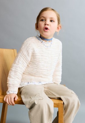 Young girl with blonde hair wearing a white sweater and beige pants sits on a wooden chair, mouth open as if speaking or singing.