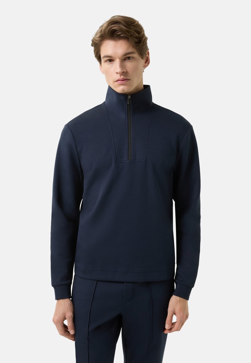 Tmavě modrý pulovr z měkkého materiálu s vysokým límcem, půlzipem vpředu a přiléhavými manžetami; doplněný o kalhoty ve stejném odstínu.