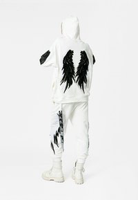 Sudadera blanca con capucha que presenta diseños de alas negras en la parte posterior y en las mangas. Combinada con pantalones a juego, también decorados con gráficos de alas.