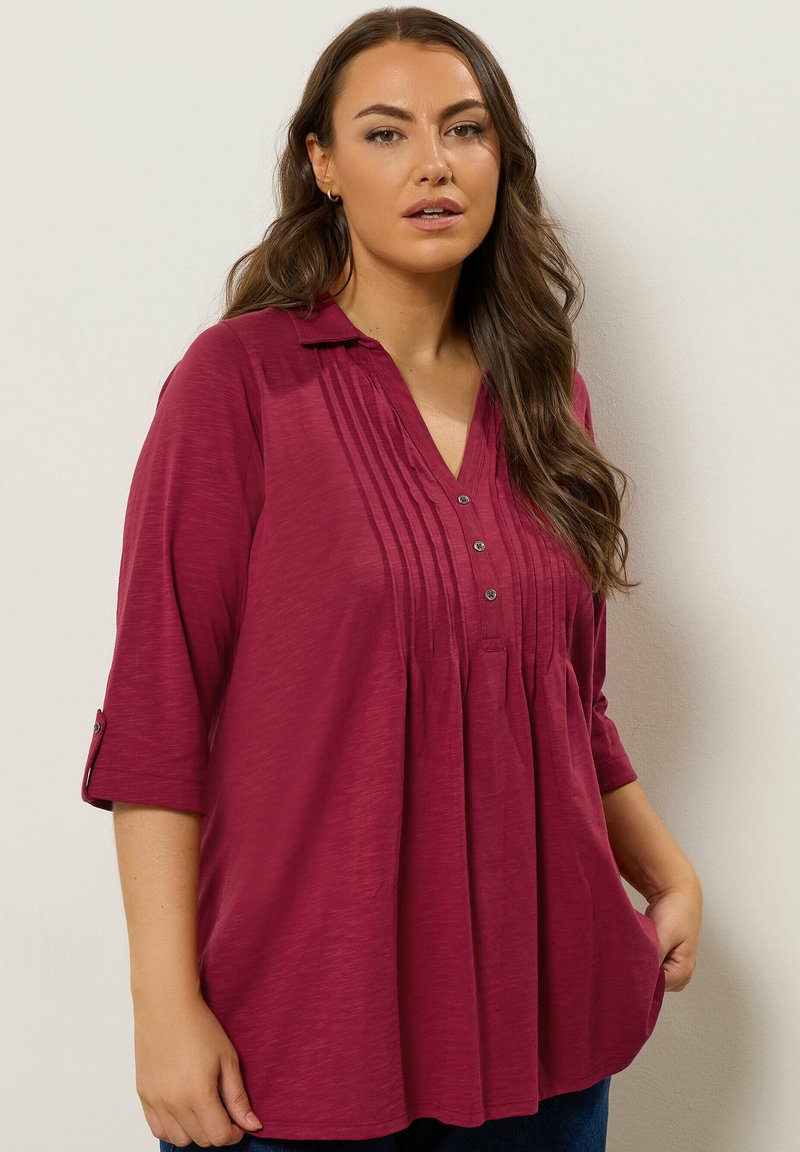 Tunic bordeaux con colletto a punta, fronte plissettato e maniche a tre quarti. Presenta dettagli con bottoni e una vestibilità ampia e fluida.