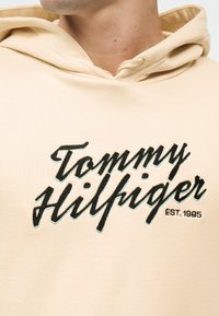Bēša kapuce džemperis ar melnu un baltu kursīvu "Tommy Hilfiger" logotipu un tekstu "EST. 1985" krūtīs, kuru valkā cilvēks.