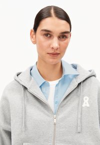 Grauer Kapuzenpullover mit Reißverschluss aus weichem Stoff, mit Kängurutasche und weißem Band-Logo. Darunter trägt die Person ein hellblaues Hemd mit Kragen.