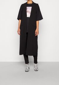 Černá dlouhá vesta bez rukávů s rovým střihem a dvěma předními kapsami, nošená přes černé tričko. Kombinováno s kotníkovými botami na vysokém podpatku se zebra vzorem.