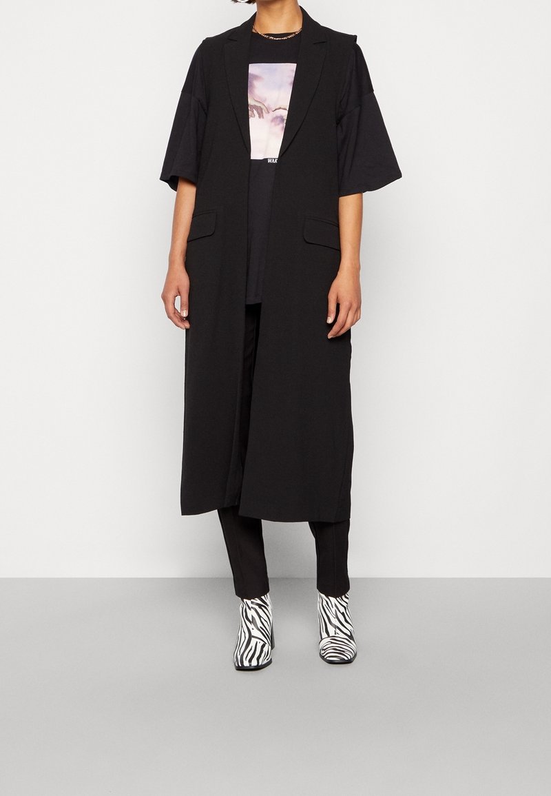 Černá dlouhá vesta bez rukávů s rovým střihem a dvěma předními kapsami, nošená přes černé tričko. Kombinováno s kotníkovými botami na vysokém podpatku se zebra vzorem.