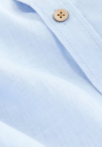 Lyseblåt linstof med en glat tekstur, der har en træknapdetalje. Stoffet er let krøllet, hvilket antyder blødhed.