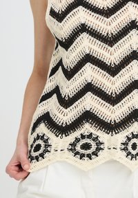 Top sans manches en crochet noir et crème avec motifs en zigzag et circulaires, porté avec un pantalon blanc, main tenant le bord du tissu.