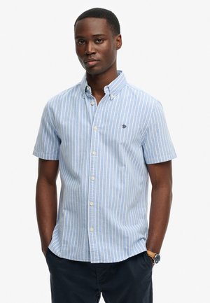 Mężczyzna stoi z rękami w kieszeniach, ubrany w jasnoniebieską i białą koszulę w paski z krótkim rękawem i ciemne spodnie, zegarek na lewej ręce.