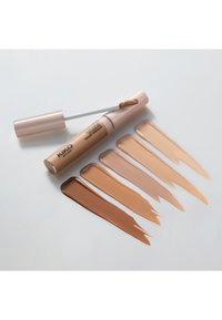 Concealer-Tube mit Applikatorstab neben fünf Mustern in verschiedenen Beige- und Brauntönen auf weißer Oberfläche.