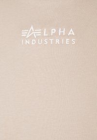 Camiseta beige con una textura suave y lisa, y un logo bordado en blanco de "ALPHA INDUSTRIES" centrado en el pecho.