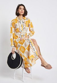 Long-sleeve button-up dress featuring a yellow and white pattern with abstract shapes and text, paired with beige slide sandals and a round black bag.
