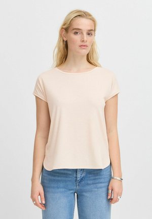 Femme aux longs cheveux blonds, portant un t-shirt beige clair à manches courtes et un jean bleu, debout devant un fond clair uni.