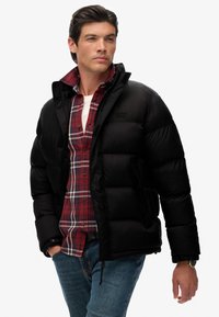 Chaqueta de plumas negra con textura acolchada, cierre frontal con cremallera, bolsillos laterales, sobre una camisa de franela con cuadros rojos, combinada con jeans azules.