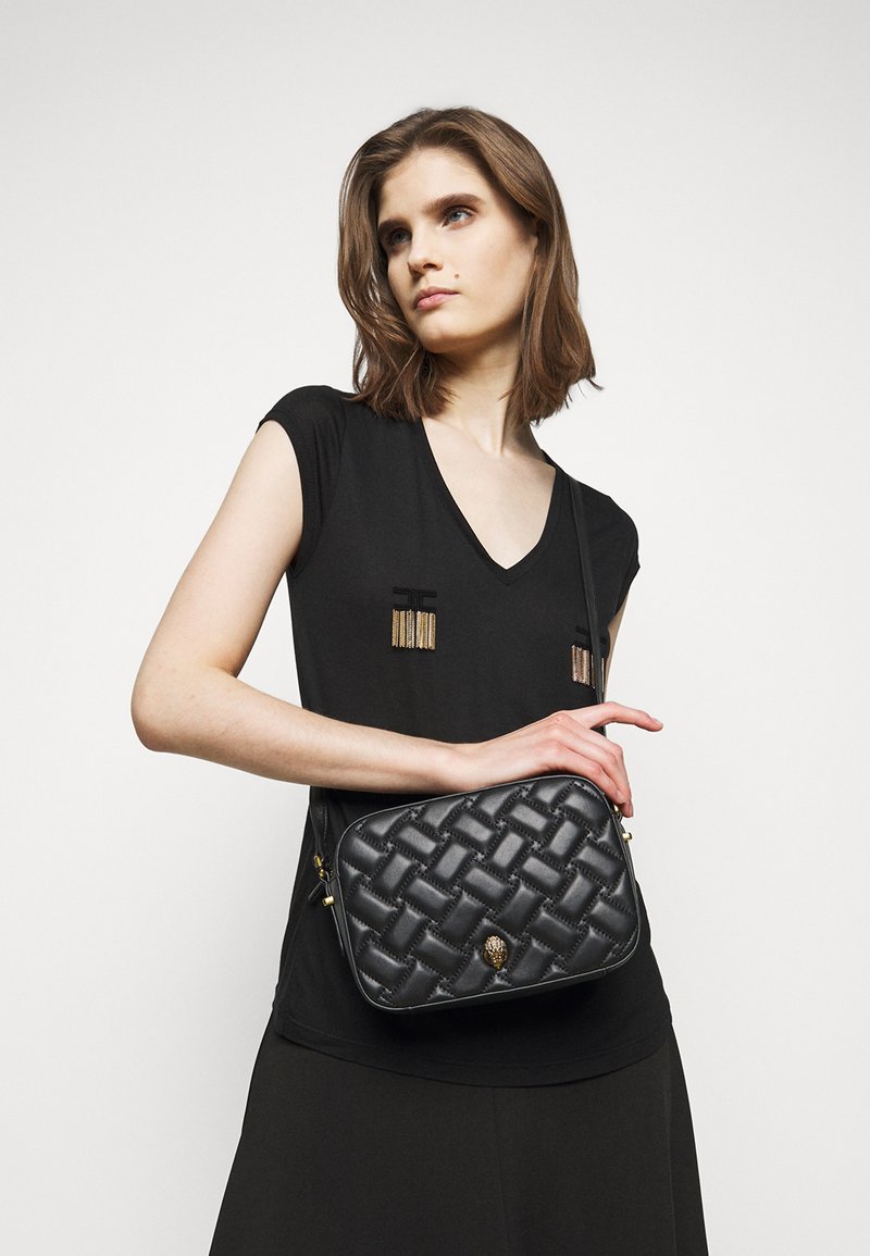 Borsa a tracolla in pelle intrecciata nera con chiusura zip e dettagli in hardware dorato, mostrata su un modello che indossa un top nero a scollo a V.