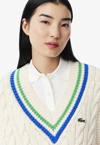 Maglione a coste color crema con scollo a V, dotato di accenti a strisce verdi, blu e bianche e un piccolo logo del coccodrillo ricamato.