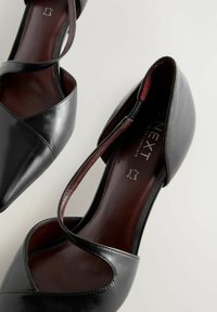 Czarne skórzane szpilki z ostrym noskiem, krzyżującymi się paskami i bordowym wnętrzem. Gładka faktura i minimalistyczny design.