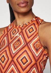 Gros plan d'une personne portant un haut sans manches orange avec un motif en losanges et un petit logo blanc sur la poitrine.