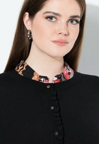 Černý cardigan s volánkovým lemem a stříbrnými knoflíky. Má živou, vzorovanou výstřih v odstínech oranžové a černé.