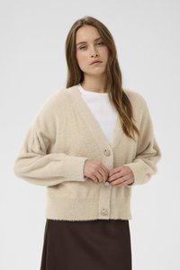 Žena s dlouhými vlasy zapíná knoflíky na béžovém huňatém svetru přes bílý top a tmavě hnědou sukni na jednobarevném pozadí.